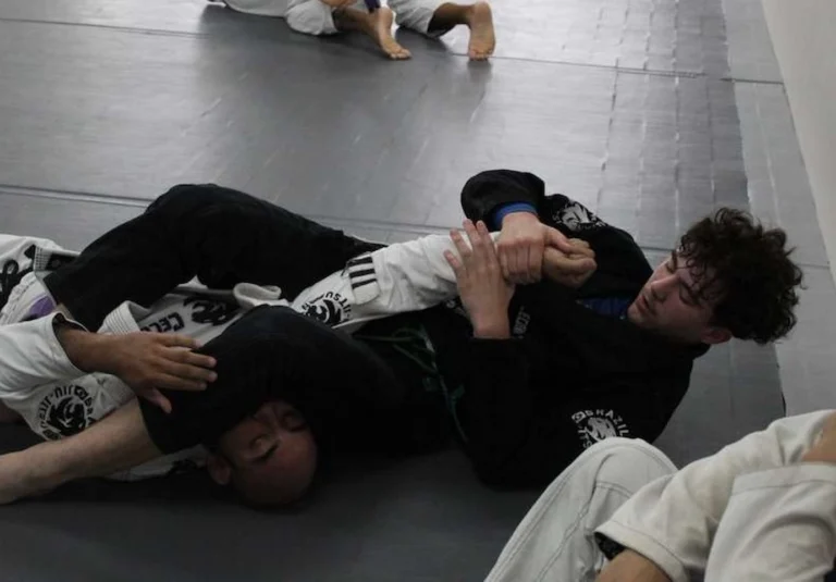 Two students in black and white gis practicing Brazilian Jiu-Jitsu on the mats. Students practicing a ground technique in the Fundamentals Jiu-Jitsu program